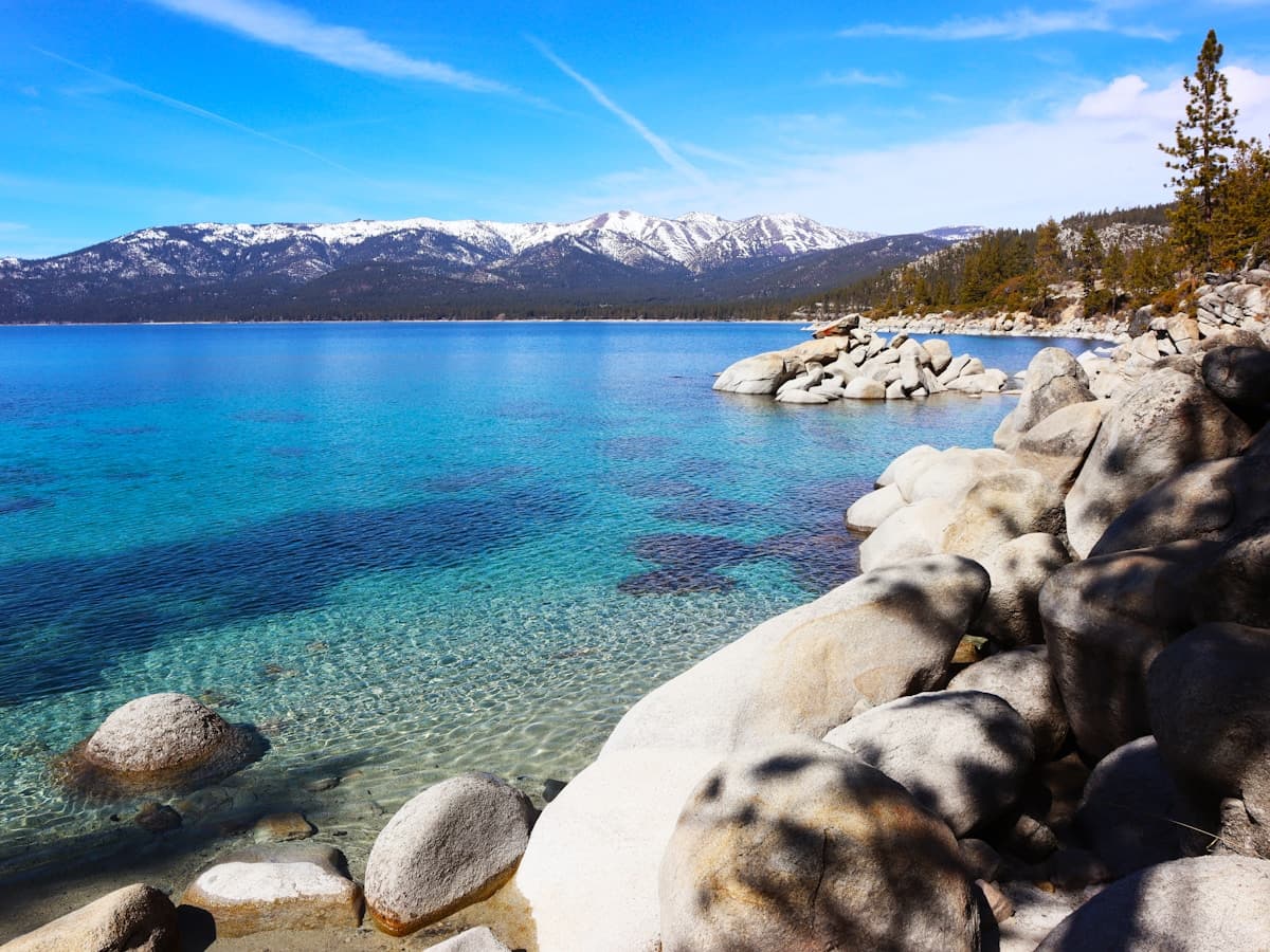 A rocky Lake Tahoe shoreline with crystal-blue water and the Sierra Nevada mountains rising on the far shore.